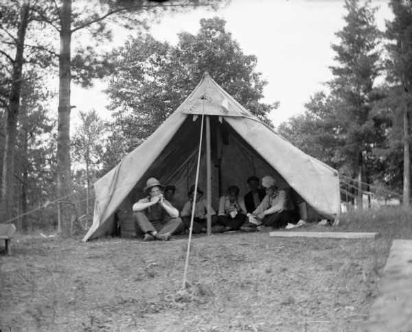 A group of young men rest inside of a tent. One man plays a harmonica while others listen and or read.