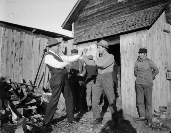 Two men take a boxing pose outside of a cabin near a woodpile. In addition, there are several onlookers.