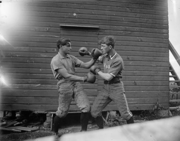 Two young men, possibly dressed in baseball uniforms, box outdoors in front of a wooden building. The man on the left is Lutie Franz and on the right is George Van Hersett.