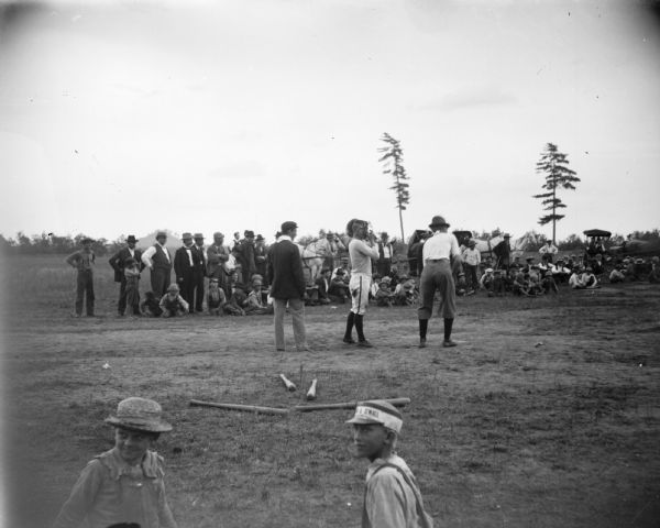 A group of spectators watch a baseball game. A batter, catcher, and umpire are visible, along with two young boys in the foreground.