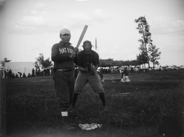 A batter for the "Hatfield" team stands at home plate with the opposing team's catcher. In the background tents and large groups of people are visible.