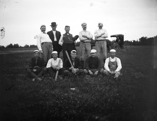 Ten men pose for a photo of their baseball team. In the background are a horse and buggy.