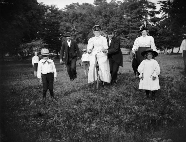 A group of men, women and children walk through a field. One woman rides a bike and is steadied by the man next to her.
