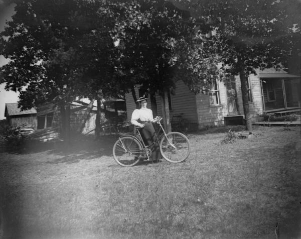 A woman stands in a yard with a bicycle. In the background, a frame house and farm buildings are visible.