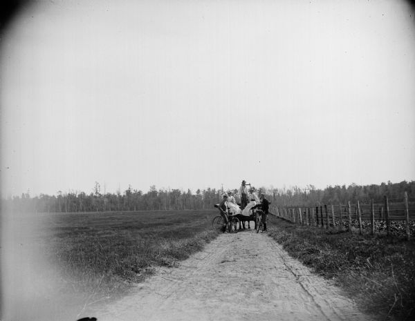 Rear view of a group of men and women with a horse-drawn wagon and bicycles stopped on a country road.