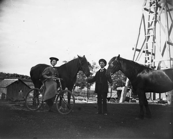 In a farmyard, a man displays two horses while a woman rests on a bike. Behind them is a tall structure with a ladder, perhaps a windmill.