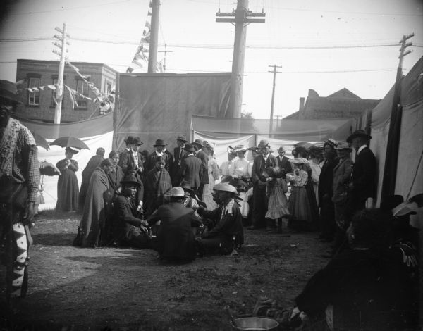 A group of Native Americans sit on the ground in a circle while other men, women, and children stand near by. There are power lines in the background.