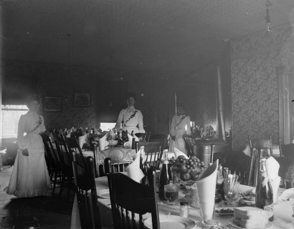 Three women pose among set banquet tables, probably at the Freeman House dining room where a meal was twenty-five cents.