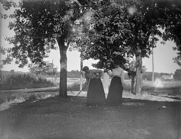 Two women play croquet in a shaded yard.