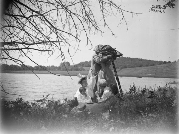 A man and a woman take a photograph on the bank of a river. In addition, there are two other women, a man, and a toddler seated on the ground near the couple.