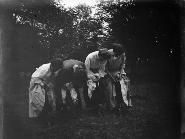 Four women are lifting the edges of their skirts up to their knees. Probably at Rock Spring Park.