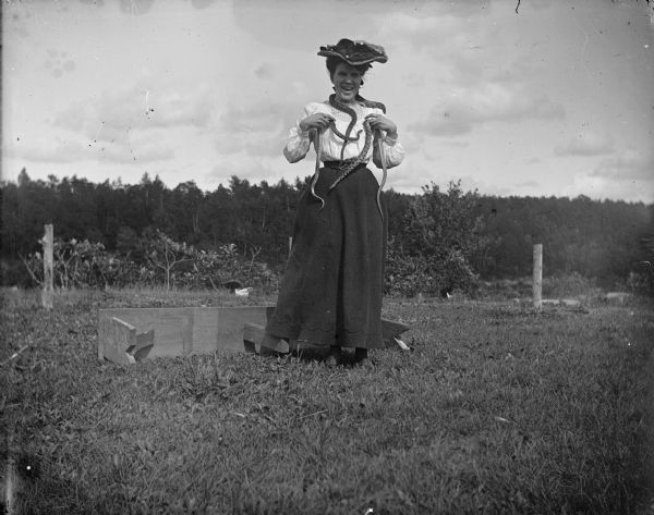 A smiling woman poses with three Pine(?) snakes around her neck and in her hands. She is standing outside, before an over-turned bench and trees, wearing a hat.