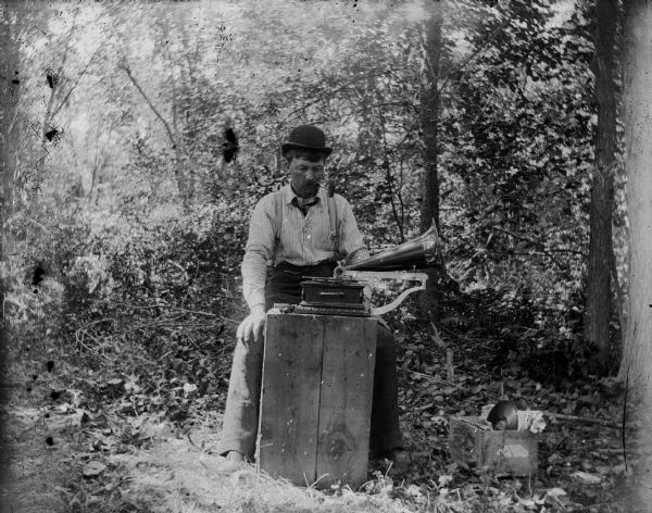 A man sits in a wooded area and plays music on a phonograph.