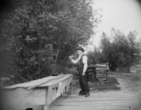A man stands near a woodpile and points a revolver toward his chest.