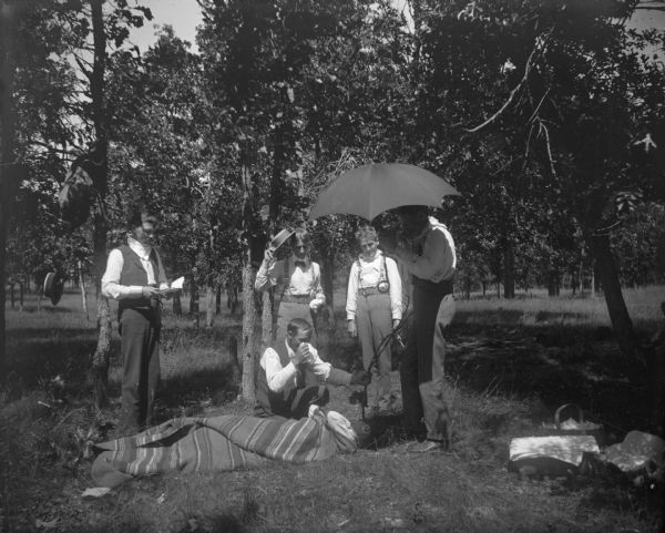 A group of men perform a mock burial at a picnic. Four men stand around a kneeling man and "corpse."	