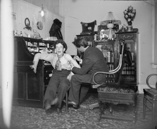 A doctor gives a patient an inoculation in an office. The two men could possibly be two medical students horse-playing. The man on the right is probably Dr. Eugene Krohn.