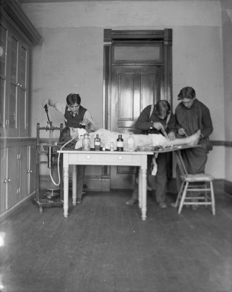 Three men, probably medical students, mimic an operation on a prostrate male. The men are identified left to right as: Price Arnold, Roy Van Schaick, Clyde Harmer, and Mahlon Richards on the table.