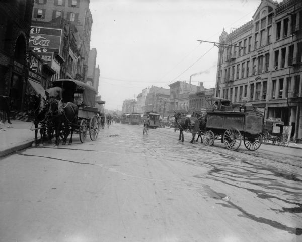 View of an urban street with pedestrians, cable cars, and horse-drawn vehicles. The city could possibly be Milwaukee, on the left there is a Schlitz Milwaukee beer sign.