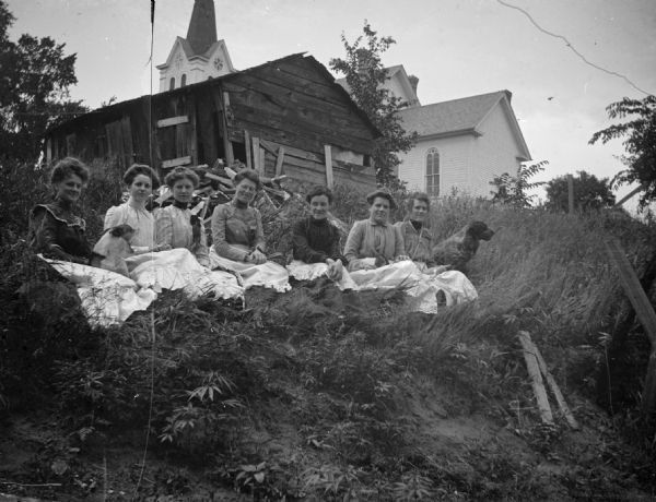 A group of seven women with two dogs sit in the grass behind the Lutheran Church.