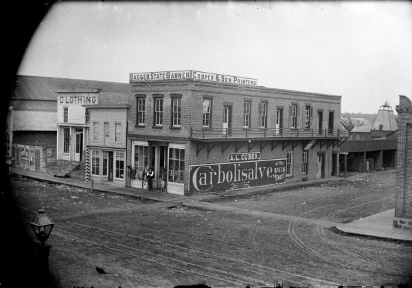 Elevated view of building of the Badger State Banner (a local newspaper) and Cooper & Son Printers. A man stands at the entrance of the building. On the side of the building is an advertisement for Carbolisalve, an ointment for the skin.