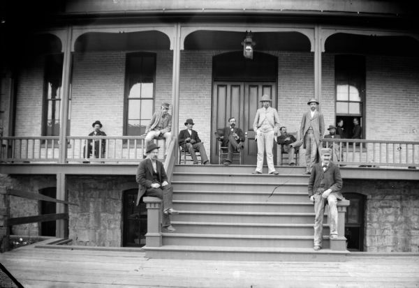 A group of men sit on the porch and steps of a brick building. In addition, two women sit on the windowsill.