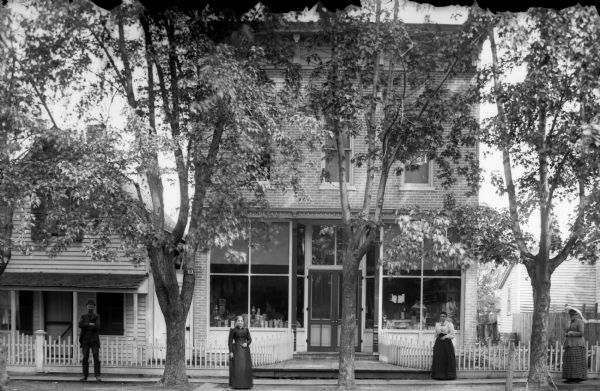 Three women and a man stand on a tree-lined, board sidewalk in front of a storefront and houses.