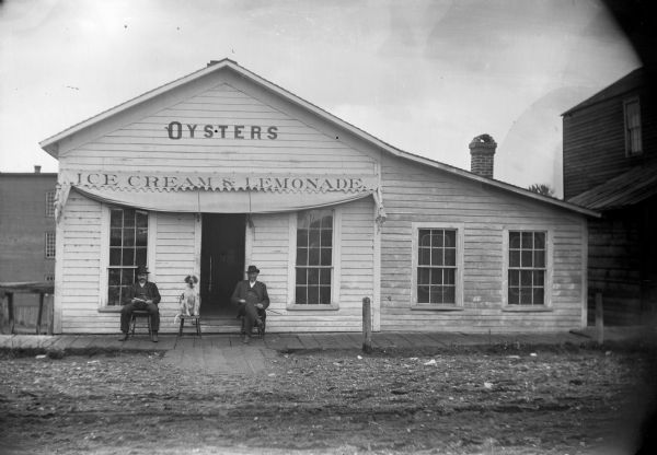 Two men and a dog sit in front of a restaurant, probably the Donnaganna Restaurant owned by R.D. Squire on Water Street. The man on the right is probably Absolom Ericson, possibly a one time cavalryman during the Civil War and later an operator of a hardware store.