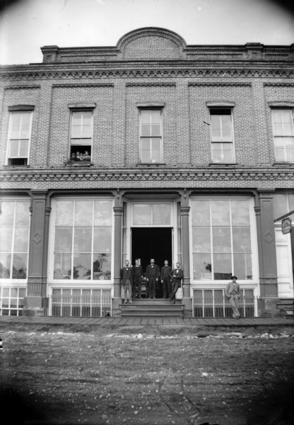 Five men stand in the doorway of the S. P. & R. C. Jones Lumber and Mercantile Company. Additionally, another man stands on the sidewalk and others look out from a second story window.