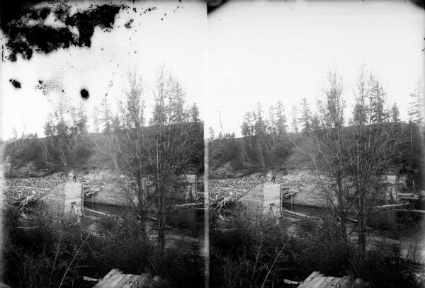 A possible stereograph of a man standing on a log jam at a sorting pier in a river. 	