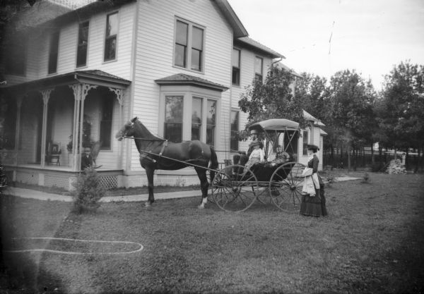 Two children and their father sit in a carriage pulled by a single horse while their mother stands to the side. The carriage is stopped in a yard of a large, two-story house.