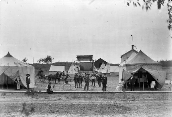 A group of people posed among tents, probably a traveling medicine show. On the table in the foreground, a contortionist is visible. A banner above a stage says, "The Umatilla Indian Hogar, for Long Life and Good Health. "