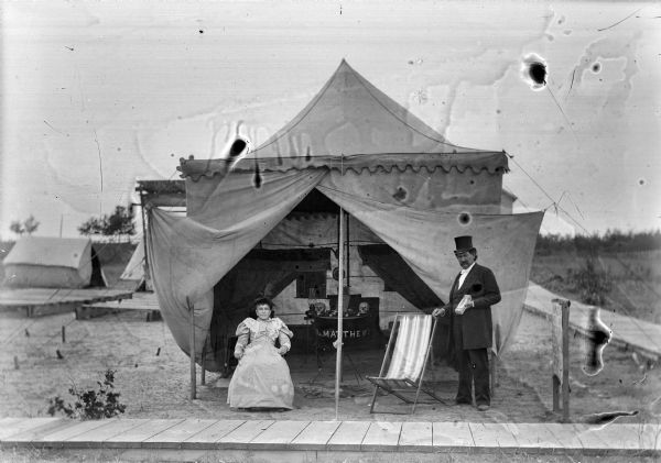A woman sits and a man stands at the opening of a tent, probably Dr. Matthews of a traveling medicine show. Inside the open tent there is a table with a display of skulls.	