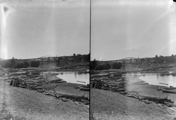 A possible stereovies of a river with logs along the shore.  Possibly the Hatfield and Railroad bridges in the background.	