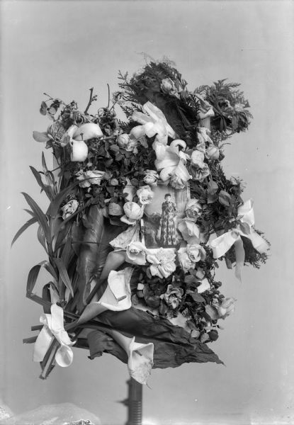 A wreath of roses and feathers surround a photograph of a studio portrait of a standing woman. 	