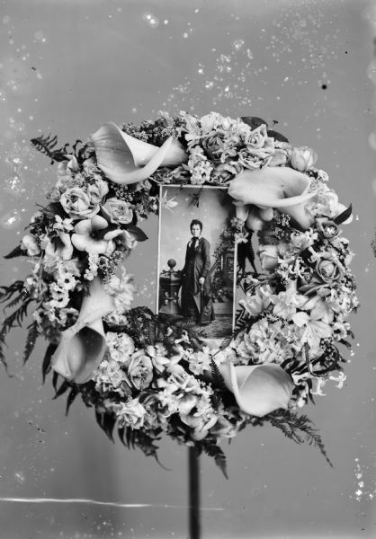 A wreath of lilies and roses surrounds a photograph of a studio portrait of a standing woman.