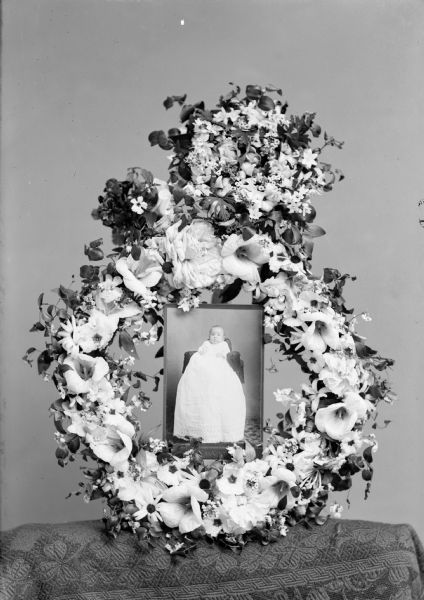 A flower wreath surrounds a studio portrait of an infant.	