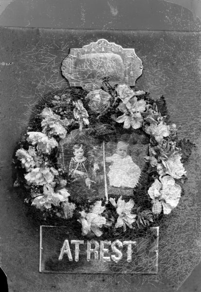 Funeral wreath surrounding two studio portraits of a boy and baby,with two attached panels "Our darling" above and "AT REST" below.  Possibly a grave stone.