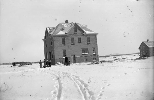 Winter scene with a man, two boys, and two girls standing in the snow outside a two-story brick building, possibly in the town of Vaudreuil.	