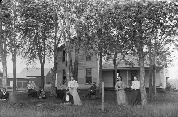 A group of men, women, and children gather in the yard of a two-story frame house.