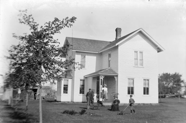 A young boy is mowing the lawn, while two women and a man are posing in the yard and on the porch of a two-story house.
