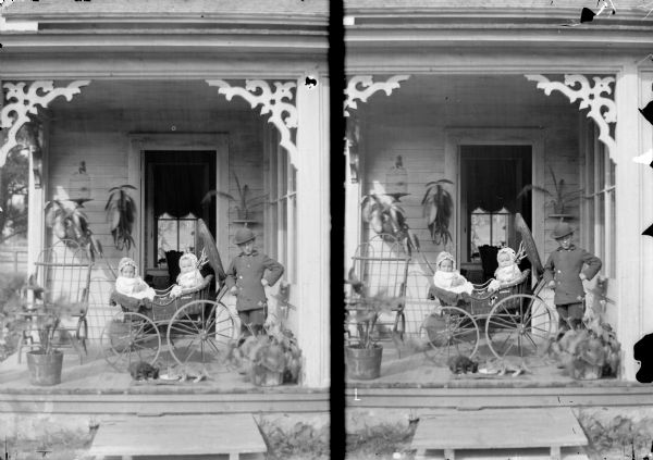 A stereograph of a boy standing next to a baby carriage with two infants sitting inside. In front of the buggy, two cats are drinking out of a saucer.