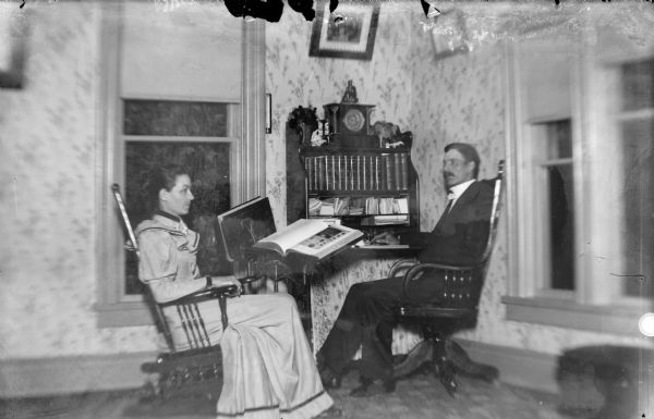 A man and a woman sit by a desk in a corner of a room that holds various books and papers.