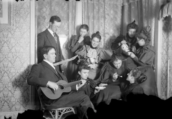 Seven women and two men gather for tea, while one of the men plays a guitar. They are posed in the corner of a room.