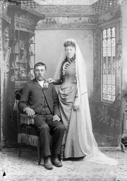 A studio wedding portrait in front of a painted backdrop. The groom is sitting in a chair in a suit jacket, vest, and trousers, with flowers in his lapel, and the bride stands with a hand on his shoulder in a dress and long veil.