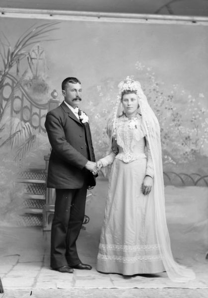 A married couple stands, holding hands, for a wedding portrait in front of a painted backdrop. He is wearing flowers in the lapel of his suit jacket, and she wears a long dress and veil.