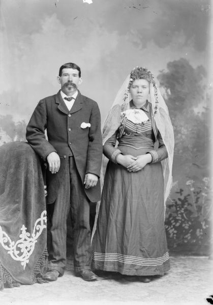 A couple, possibly Gust Schultz and Ida Gehring, poses for a wedding portrait in front of a painted backdrop. She is wearing a long dress with a veil, and he wears a knee-length suit jacket with vest and trousers.