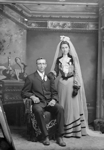 A bride and groom pose for a wedding portrait in a photography studio in front of a painted backdrop. The groom sits in a chair with flowres in his lapel, while the bride wearing a long dress, veil, and dark gloves stands and rests her hand on the groom's shoulder.
