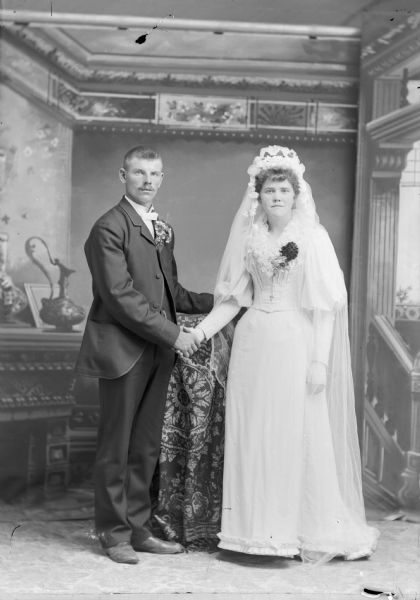 A bride and groom stand, holding hands, for a studio wedding portrait in front of a painted backdrop. He is wearing flowers in his lapel, and she is wearing a long dress and veil.