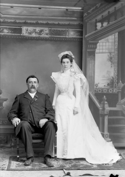 A bride and groom pose for a studio wedding portrait in front of a painted backdrop. The groom sits in a chair with flowers in his lapel, as the bride in his long dress and veil stands to his left side.