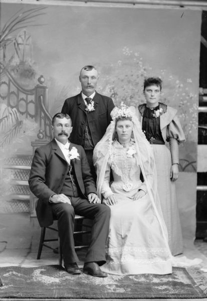 A bride and groom sit while two attendants stand behind them for a studio portrait in front of a painted backdrop. The bride wears a long dress and veil, and the groom wears flowers in his lapel.